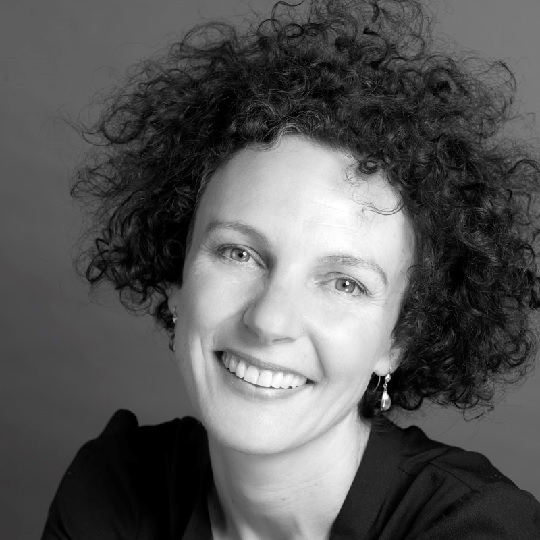 Black and white photo of Hilary Bell, Hilary has dark curly short hair, she is smiling and her teeth show. She has little drop earrings and wears a dark coloured shirt